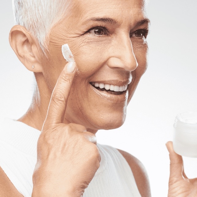 Glimlachende rijpe vrouw die anti-aging crème op haar gezicht aanbrengt.