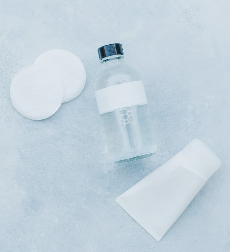 Een fles gezichtsreiniger, twee wattenschijfjes en een tube crème op een lichte achtergrond, symbool voor huidverzorgingsproducten.